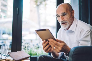 Olgun, kel bir adamın resmi kıyafetler giymesine şaşırdım. Modern tableti karıştırıyor. Kafenin masasında şaşkın şaşkın oturuyor.