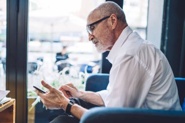 Kafedeki mavi rahat koltukta oturan odaklanmış ve konsantre olmuş iş adamının yan görüntüsü. Akıllı telefon ve sosyal ağda mesajlaşma.