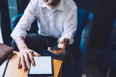 İsimsiz gri sakallı, beyaz tişörtlü yaşlı bir adam mavi sandalyede oturuyor ve tablet üzerinde gezinirken akıllı telefon kullanıyor.