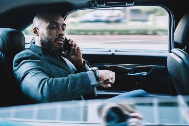 Kendine güveni tam, kel ve sakallı siyah iş adamı. Zarif giysiler içinde cep telefonuyla konuşuyor ve arabada oturmuş saatine bakıyor.