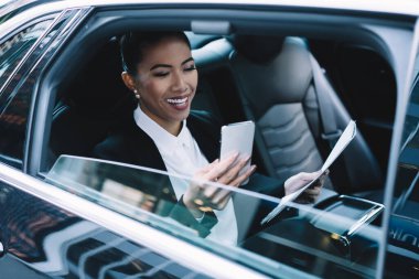 Resmi olarak giyinen parlak etnik bir bayanın arabasının arka koltuğunda oturan telefondaki mesajları analiz etmesi.