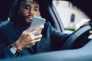 Kol saati ekrana odaklanan ve trafik sıkışıklığında araba kullanırken akıllı telefonla etkileşen ciddi Afrika kökenli Amerikalı iyi giyimli iş adamından.