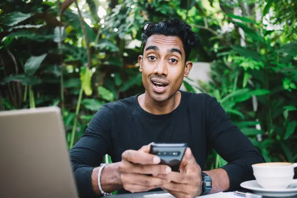 Shocked adult Hispanic guy with opened mouth in black shirt looking at camera while sitting at table with laptop papers and hot drink and interacting with smartphone in garden on blurred background of big green plants