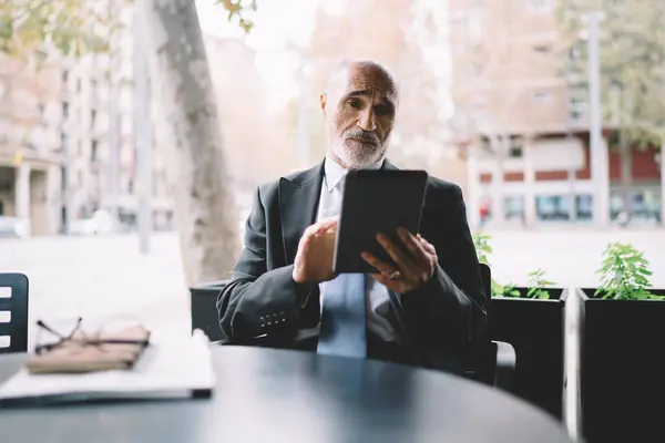 Pensive elderly businessman in classic suit sitting in cafe in front of papers and glasses on table while messaging in social network