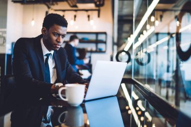 Modern kafeteryanın tezgahında bir fincan kahveyle otururken laptopla çalışan ve haberleri okuyan ciddi bir konsantre olmuş Afro-Amerikan işadamı.