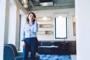 Düşünceli, sorunlu, resmi giysiler içinde Asyalı bir kadın lobideki kanepenin önünde dikilirken ve yan yatarken telefon görüşmesi yapıyor.