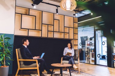 Ciddi iş adamı dizüstü bilgisayar kullanıyor ve tabletli genç Asyalı kadın ofis bekleme odasında oturmuş iş toplantısına hazırlanıyor.