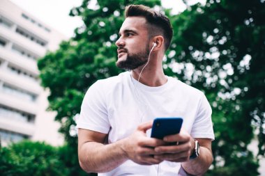 Kulaklığı ve akıllı telefonu olan neşeli ve iyimser bir adamın park yerinde durup sosyal medyada mesajlaşırken gülümsemesi ve başka yere bakması.