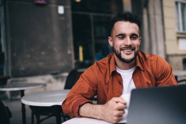 Neşeli, yakışıklı, sakallı, sakallı adam yazın, modern kafeteryada sıcak bir bardak sıcak içecek içerken zevkle sörf yapıyor.