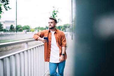 Sakallı, sıradan kıyafetli genç ve yakışıklı bir hippi sokakta akıllı telefonuyla dikilirken düşünceli bir şekilde uzaklaşıyor. 