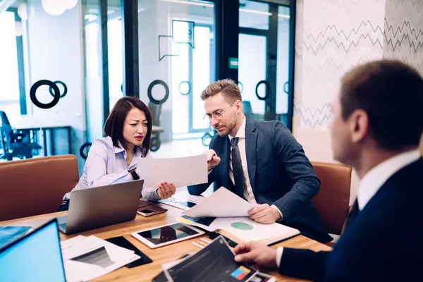 stock image Multiracial colleagues sitting at table with laptop tablet and documents and brainstorming and analyzing project in comfortable workspace of company