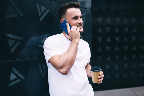 Smiling bearded male in casual clothing talking on smartphone and holding disposable coffee cup while standing in front of modern office building