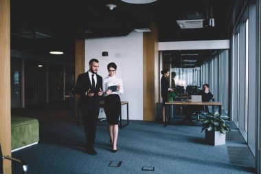 Resmi kıyafetli bir adam ofis koridorunda beklerken elinde tabletle gözlüklü güzel bir kadınla konuşuyor.