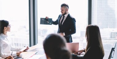 Modern ofis binasının penceresinin yanında dururken şık saç kesimi olan sakallı bir yönetici yönetim kuruluna tabloyu sunuyor.