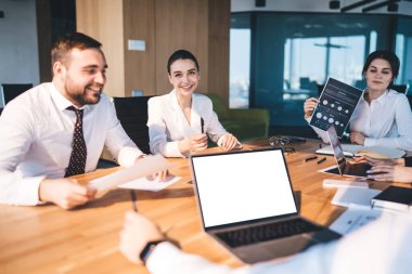 Resmi olarak gülümseyen ofis çalışanları modern şehir ofisindeki belgelerle konferans masasında otururken tartışma çizelgesi ve dizüstü bilgisayarda geziniyorlar.