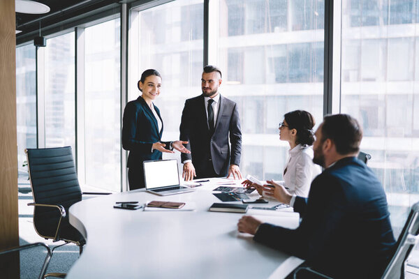 Side view of pleased adult business people in elegant clothes having discussion while looking at each other at desk near big window in modern office of skyscraper