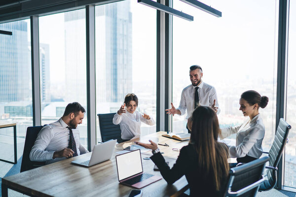 Положительные сотрудники в белых футболках и черных костюмах сидят за столом с ноутбуками и обсуждают успешный бизнес-проект в современном стеклянном офисе