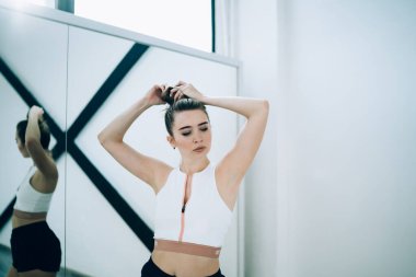 Kendine güvenen, fermuarlı, siyah şortlu, kendine güveni tam bir kadın. Beyaz, şık bir stüdyoda siyah haçı aynaya yansıtıyor.