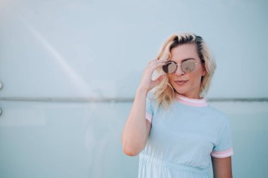 Güneş gözlüklü, şık saçlı moda kadını güneşli bir günde mavi duvarın arka planında güneş gözlüklerine dokunarak kameraya bakıyor. 