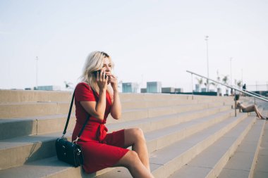 Sarışın, kısa kırmızı elbiseli bayanın akşam vakti büyük taş merdivenlerde oturup akıllı telefonu iki eliyle tutarken konuşmasının yan görüntüsü.