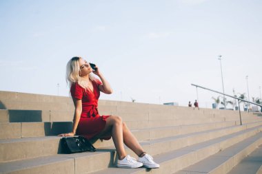 İçi rahat kırmızı elbiseli sarışın şehir merdivenlerinde dinlenirken cep telefonundan arıyor.