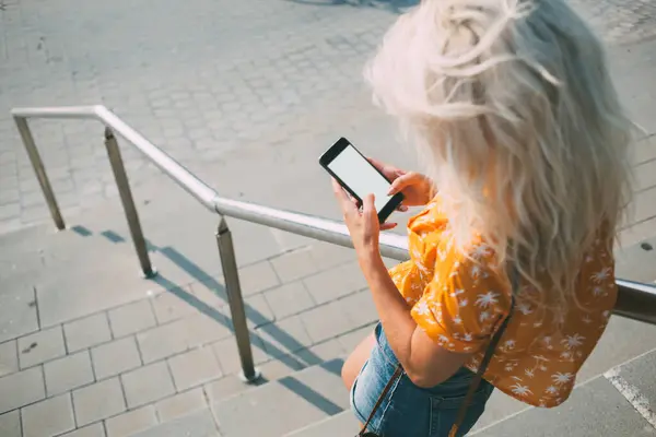 From above anonymous woman in casual outfit standing near railing on stairs and messaging on smartphone with blank screen on sunny day in city