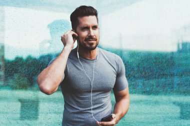 Spor elbiseli, kulaklık takan, cep telefonuyla müzik dinleyen, sokakta koşuya hazırlanan ve gözlerini kaçıran güçlü bir adam.