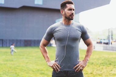 Spor kıyafetleri içinde elleri belinde, kulaklıklı, müzik dinleyen spor antrenmanlarına hazırlanan, sokakta dikilip gözlerini çeviren, kendinden emin kaslı bir adam.