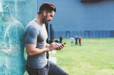 Spor kıyafetli, kulaklıklı, cep telefonlu, müzik dinleyen, stadyumdaki camdan duvara yaslanmış koşuya hazırlanan ciddi kaslı erkeklerin yan görüntüsü.