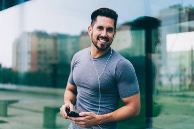 Spor kıyafetleri içinde, kulaklıklı, telefon kullanarak sokakta koşuya hazırlanan müziği dinleyip kameraya bakan neşeli bir erkek.