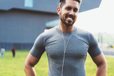Spor kıyafetli, kulaklıklı, müzik dinleyen, spor egzersizi hazırlayan ve sokakta kameraya bakan mutlu, kaslı adam.