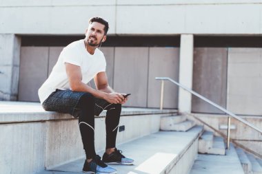 Spor kıyafetli, kulaklıklı, cep telefonlu, cep telefonlu, merdivenlerde oturmuş müzik mesajı dinleyen düşünceli yakışıklı bir erkek.