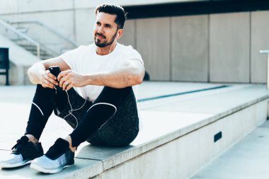 Şık etkin sporcu tatmin olmuş erkek sporcu merdivenlerde oturuyor ve gözlerini kaçırıyor. Şehir caddesinde cep telefonu ve kulaklıkla müzik dinliyor. 