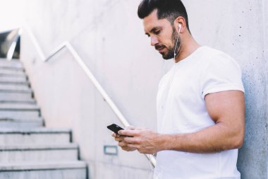 Düşük açılı, siyah sakallı, beyaz tişörtlü, kulaklıkla gezen ve şehir caddesindeki binanın duvarına yaslanan genç adam. 