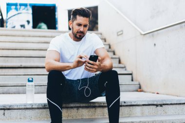 Aktif görevde olan sakallı erkek sporcu, antremandan sonra sallanan merdivenlerde otururken kulaklıkla cep telefonuyla haberleri kontrol ediyor. 