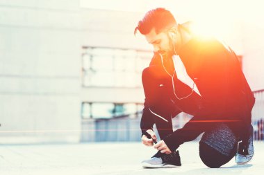 Koyu renk spor giyimli güçlü bir koşucunun yan görünüşü kulaklıklarla diz çöküp ayakkabı bağcıklarını bağlarken müzik dinleyip, parlak bir günde koşması.