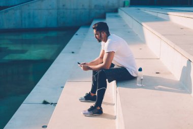 Spor kıyafetli, kulaklıklı, egzersiz sonrası dinlenen telefonda konuşan, merdivenlerde oturan aktif bir adamın yan görüntüsü.