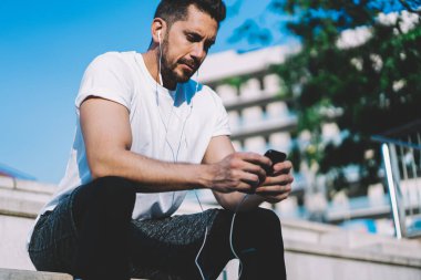 Düşünceli, sakallı, kulaklıklı, cep telefonlu ve parlak bir günde merdivenlerde oturmuş müzik dinleyen bir sporcu. 