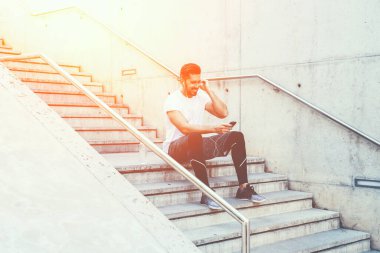 Spor kıyafetleri içinde, kulaklıkları ayarlayan ve merdivenlerde otururken ve şehirde eğitime hazırlanırken cep telefonundan çalma listesini seçen neşeli ve aktif bir erkek.