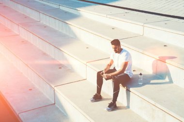 Spor adamının cep telefonu kullandığına ve kulaklıkla müzik dinlediğine yukarıdan bakıldığında güneşli bir günde bir şişe suyla merdivenlerde otururken ve dinlenirken görülüyor.