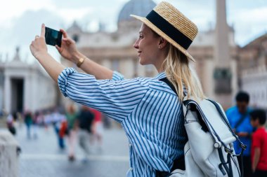 Moda kıyafetleri içinde çekici bir kadın modern cep telefonu kamerasıyla resim çekiyor. Yüksek kalitede fotoğraf çekiyor, kaygısız turist Vatikan 'da İtalyan tatili hakkında video bloğu çekiyor.