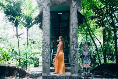 Uzun turuncu elbiseli, çekici, uzun saçlı bir kadın. Bali tropikal ormanlarında Budist heykeliyle taş köprü üzerinde duran bacağını gösteriyor. 