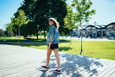 Kot ceketli, siyah şortlu, gülümseyen genç bir kadının şehir parkında yürürken kameraya baktığı ve güneşli yaz gününün tadını çıkardığı yan manzara.