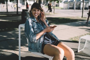 Düşük açılı, kot ceketli, siyah şortlu, kulaklıklı, akıllı telefon kullanırken kameraya gülümseyen ve boş zamanlarında sokakta müzik dinleyen bir öğrenci.