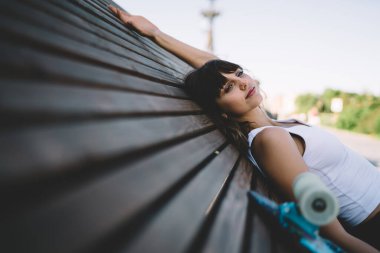 Beyaz tişörtlü, kaykayın yanındaki büyük ahşap bankta oturan ve güzel bir yaz gününün tadını çıkaran genç aktif bir kadının yan görüntüsü. 