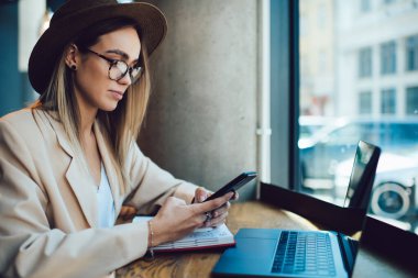 Gözlüklü, şapkalı, ekranda odaklanan ve modern binanın penceresinin yanındaki bilgisayarda akıllı telefon kullanan düşünceli, zarif yetişkin bir kadının yan görüntüsü.