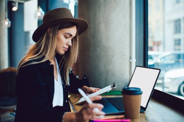 Kafedeki boş ekranlı dizüstü bilgisayarla masada otururken elinde kalem tutan ve panodan veri okuyan konsantre bir kadının yan görüntüsü