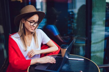 Modern şapkalı ve gözlüklü modaya uygun gülümseyen serbest yazar şehir kafeteryasındaki laptopta daktilo etmek için kahve içer.