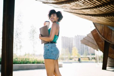 Gülümseyen, kıvırcık saçlı, yeşil tişörtlü ve şortlu mutlu etnik bayanın elinde bir fincan kahveyle sokakta yürümesinden.