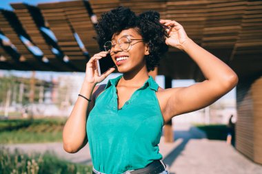 Yeşil tişörtlü, gözlüklü, kıvırcık saçlı, mutlu bir etnik kadın yazın sokakta cep telefonuyla konuşuyor.